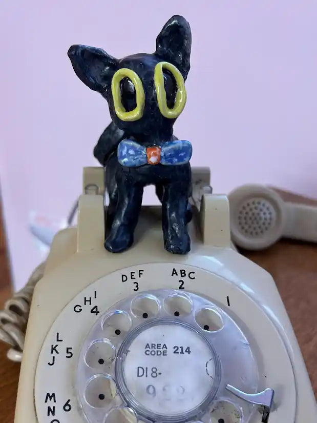 Small black cat figurine with yellow eyes and a blue bow tie sitting on an old-fashioned beige rotary telephone.