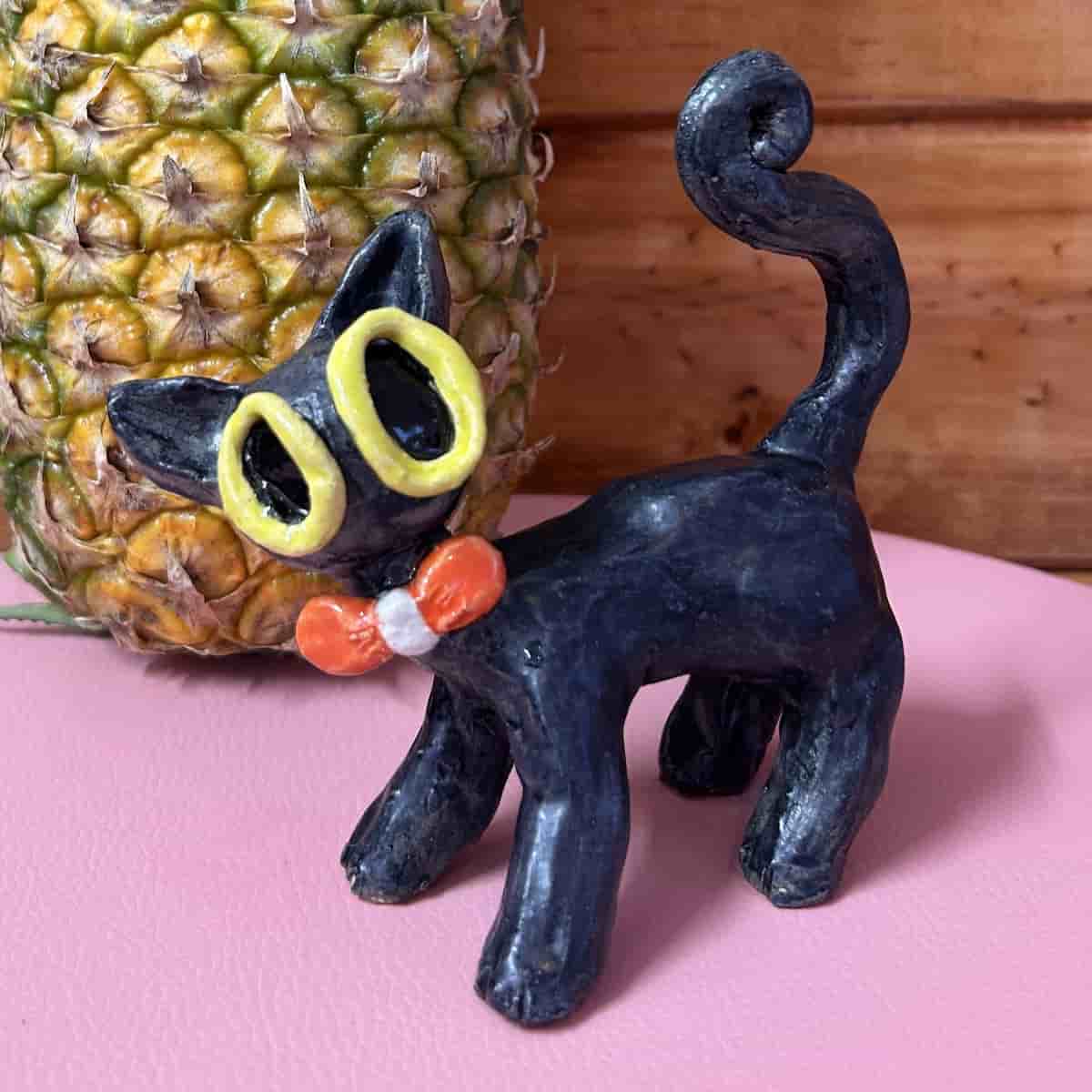 Ceramic black cat figurine with red bow tie and comical yellow eyes, standing in front of pineapple
